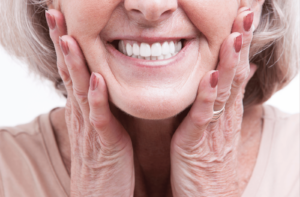 A close up image of the bottom half of a woman face, she is holding both of her hands up to her cheeks and smiling, showing her dental bridge