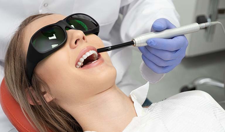 A woman sitting back in a dental chair at the dentist's office and she is receiving Solea Laser treatment by a dentist