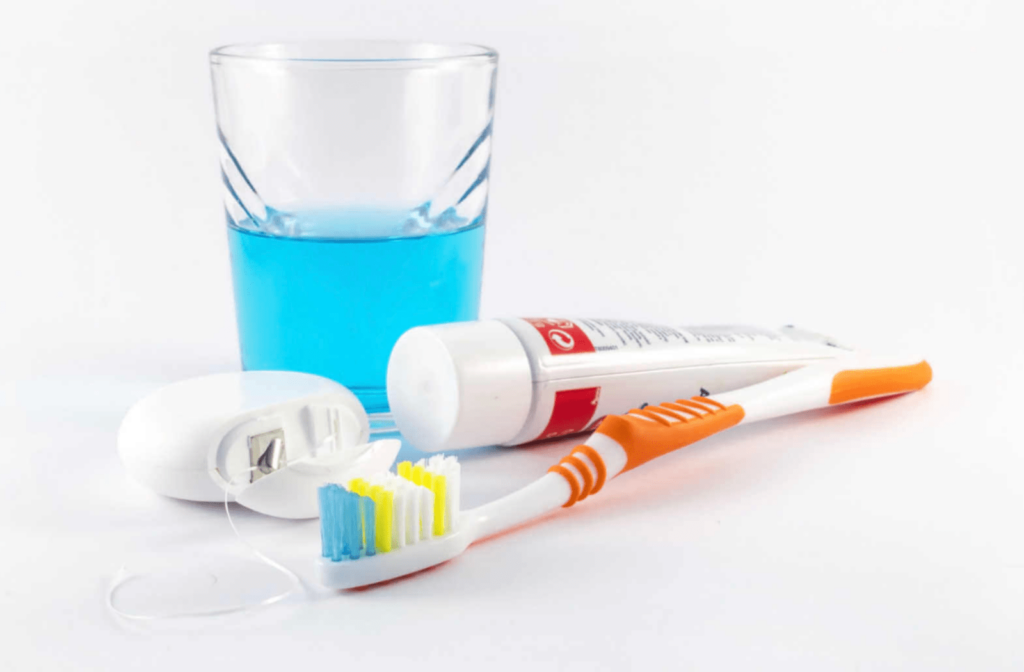 A close-up of materials for cleaning dentures, including a soft-bristle toothbrush, a tube of toothpaste, a roll of dental floss, and a half glass of mouthwash.