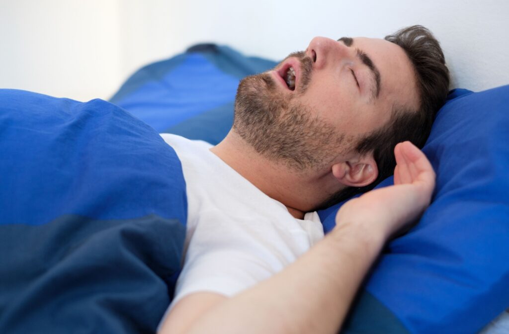 A man lying in bed on his back, covered by a blue sheet. His mouth is open as he snores