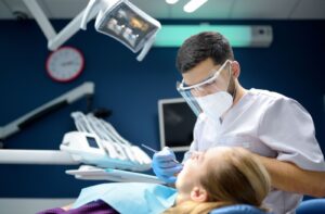 A dentist examines a patient's mouth in a modern clinical setting using a scraper while wearing a face mask for protection.