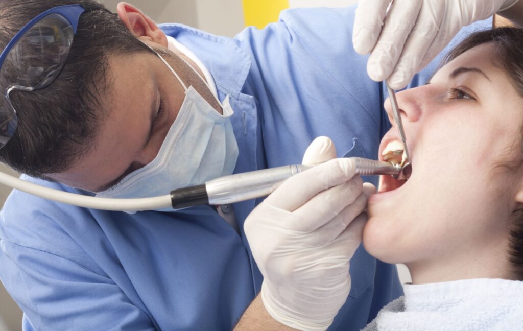 A dental hygienist is carefully cleaning their patient's teeth as the patient sits quietly with their mouth open.