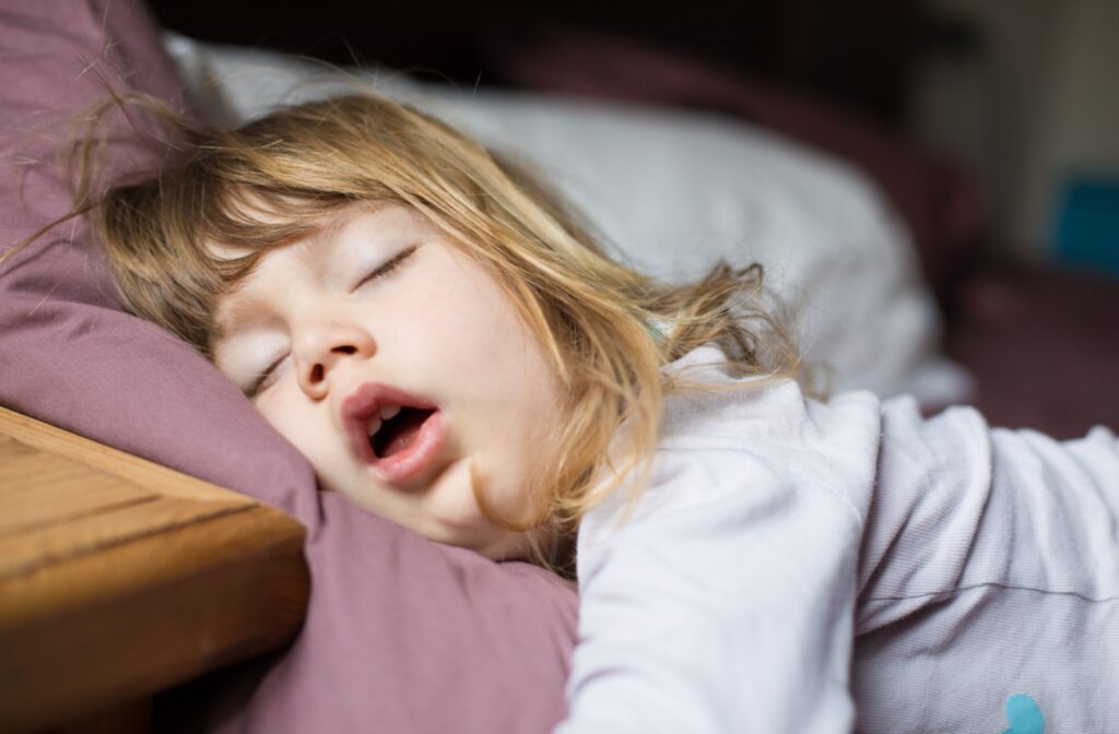 A young child lies sleeping in their bed. Their mouth is open and their head is tipped back as they snore loudly.